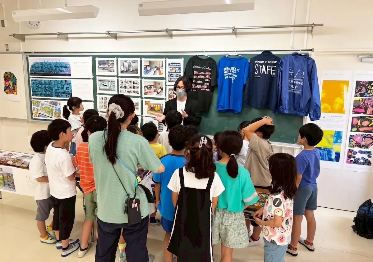 黒板前に展示されたアール・ブリュット作品を背に、当社男性社員が学生たちに囲まれながら説明している様子の写真