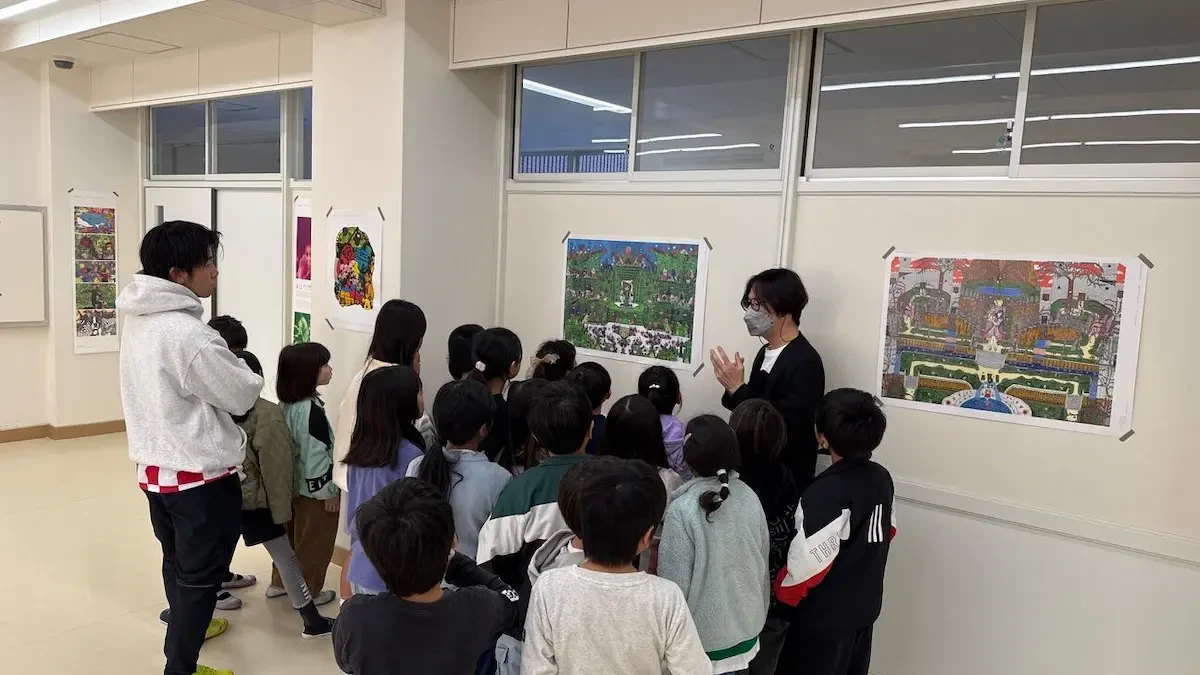 教室にさまざまなアール・ブリュット作品が展示されており、学生たちの前でアール・ブリュットについて説明している当社男性社員の写真