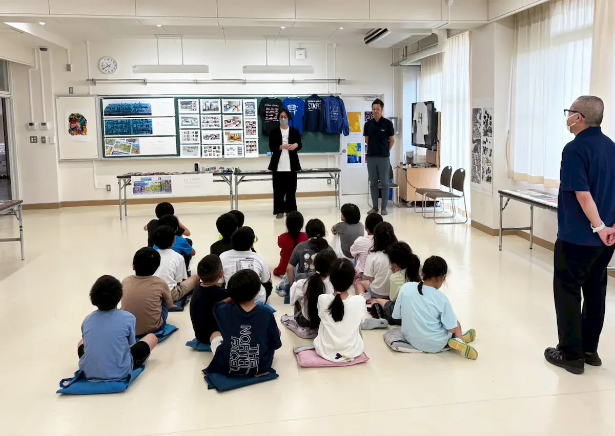 教室にさまざまなアール・ブリュット作品が展示されており、多摩市立聖ヶ丘小学校の学生たちの前でアール・ブリュットについて説明している様子の写真