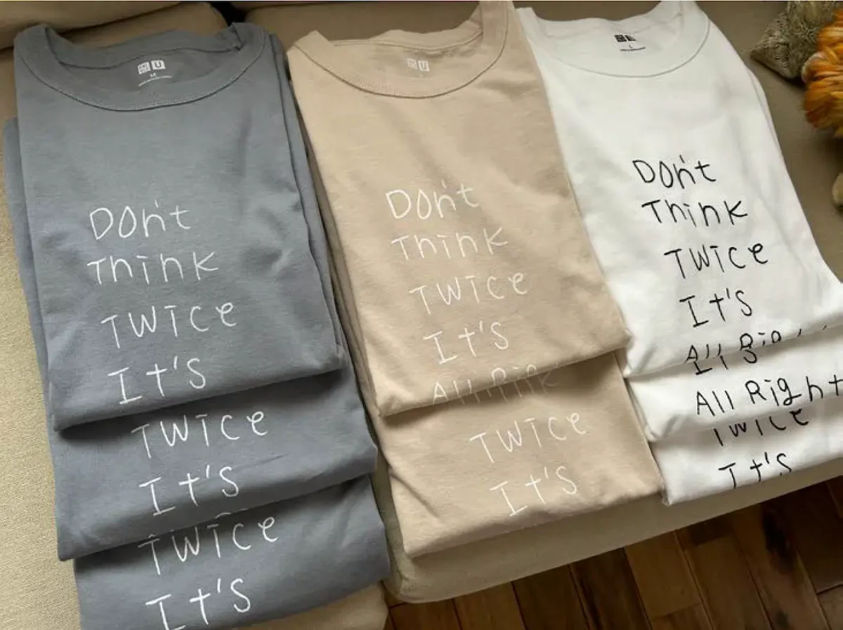 伊山英吾さんの作品がデザインされたTシャツの写真。グレー・ベージュ・ホワイトの3色のTシャツが畳まれた状態で映っています。